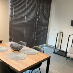 Brązowe poziome osłony okienne w nowoczesnym wnętrzu jadalni z drewnianym stołem i czarnymi krzesłami.