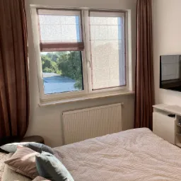 Sypialnia z dwoma oknami wyposażonymi w brązowe rolety rzymskie, brązowe zasłony po bokach okien, jasna pościel na łóżku i dekoracyjne poduszki.