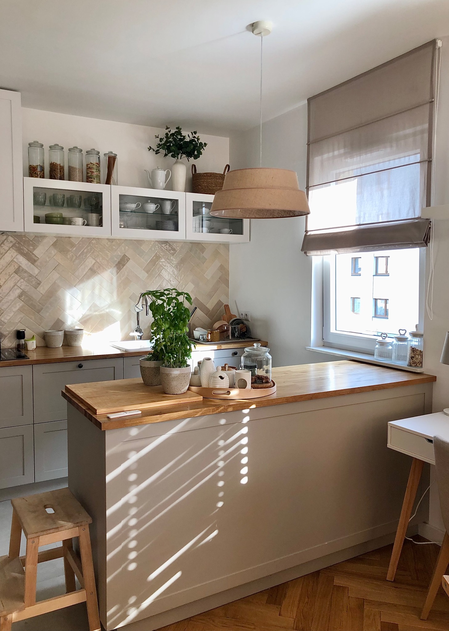 Beżowa, rzymska osłona okienna w jasnej kuchni z drewnianym blatem i dekoracyjnymi pojemnikami na półkach.