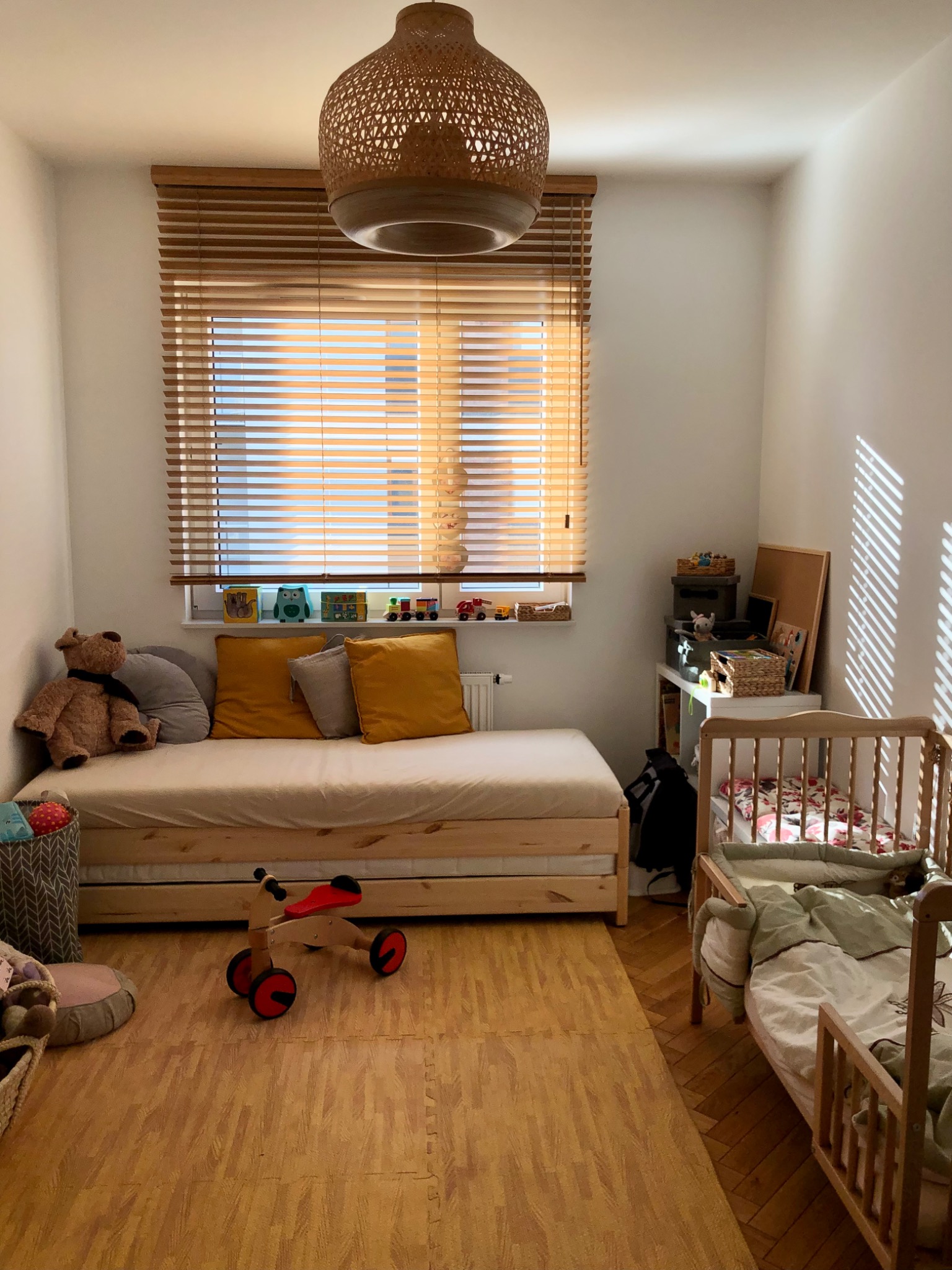 Pokój dziecięcy z drewnianymi żaluzjami poziomymi, przez które wpada ciepłe światło słoneczne, oświetlając drewniany rowerek i łóżko z pluszowym misiem
