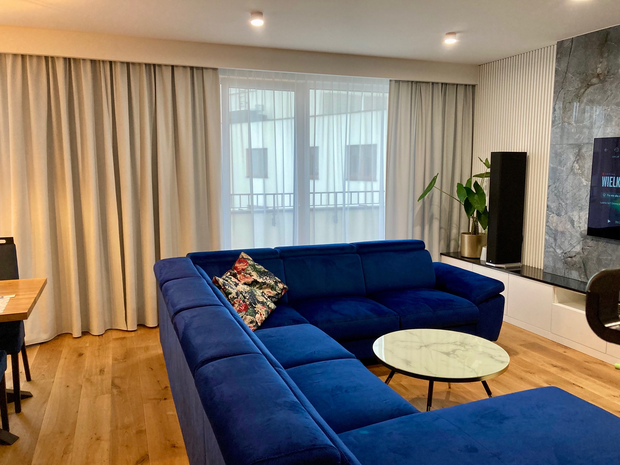 Eleganckie beżowe zasłony panelowe w nowoczesnym salonie z niebieską sofą narożną i stolikiem kawowym z marmurowym blatem.