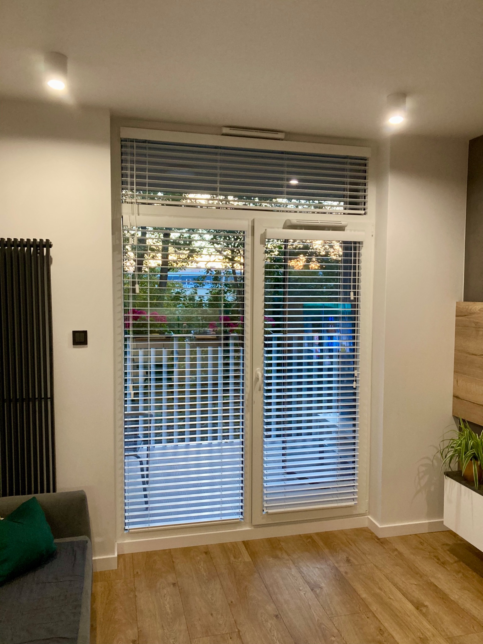 Nowoczesne wnętrze z białymi, poziomymi osłonami okiennymi zamontowanymi na dwuskrzydłowych drzwiach balkonowych, widok na ogród z zewnątrz.