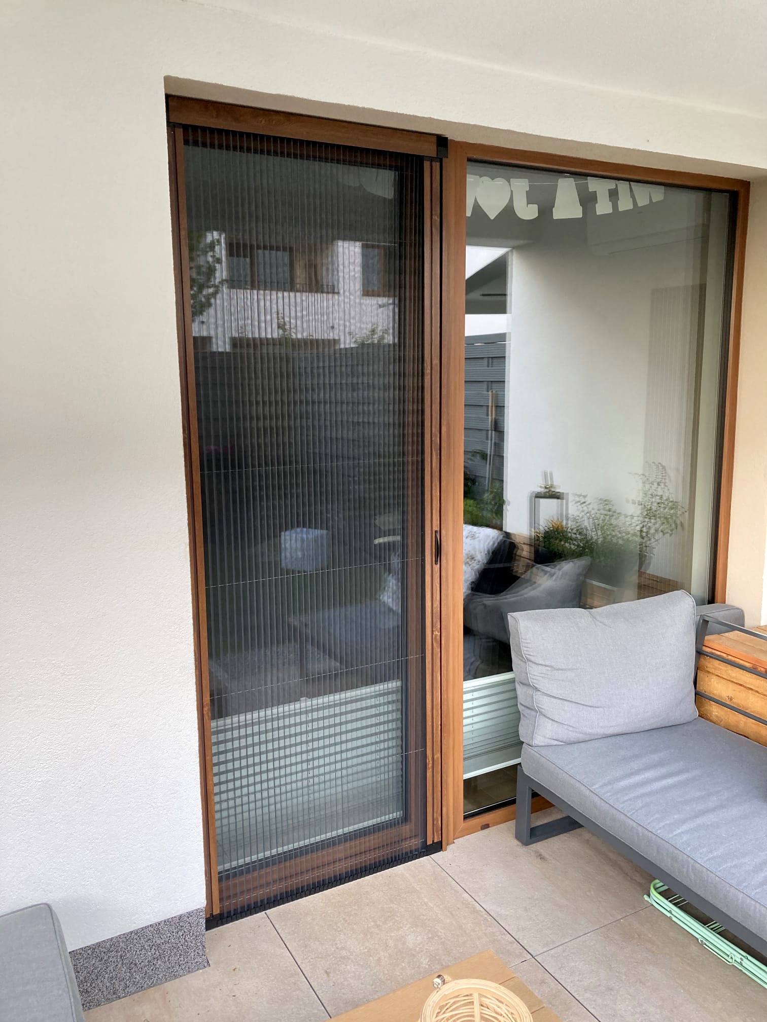 Zewnętrzna, składana przesłona okienna z drewnianą ramą, widoczna z zewnątrz, z częściowo zasłoniętym widokiem na wnętrze balkonu z szarą sofą i dekoracjami okiennymi.