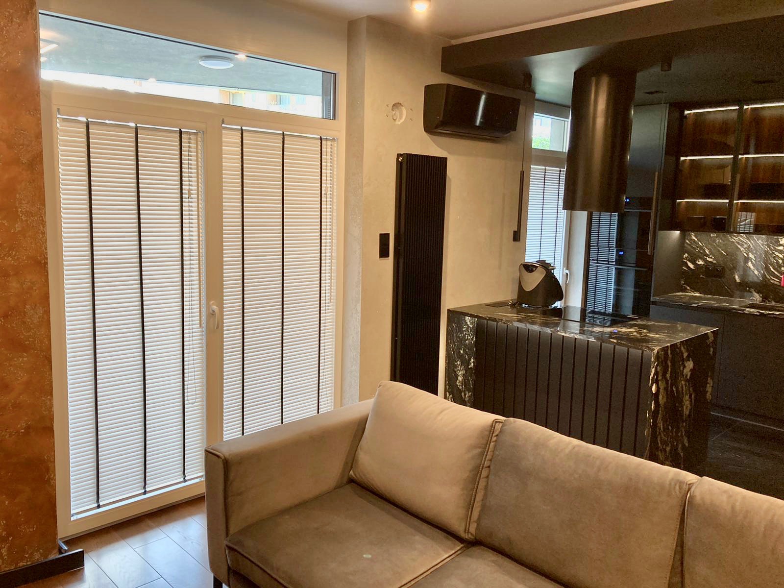 Nowoczesne wnętrze salonu z widokiem na okno balkonowe zabezpieczone białymi żaluzjami poziomymi z czarnymi sznurkami, obok beżowa sofa i wyspa kuchenna z czarnego marmuru.