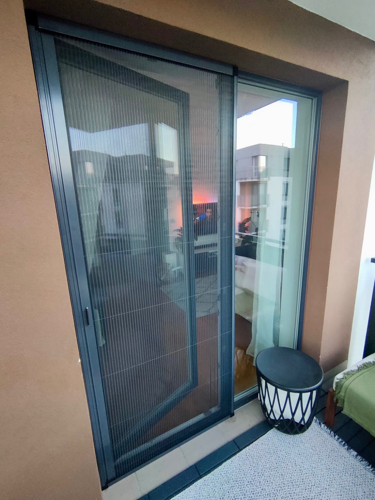 Złożona plisa okienna w ciemnej ramie na balkonie, widok na wnętrze mieszkania z włączonym telewizorem i widok na zewnątrz na inny budynek.