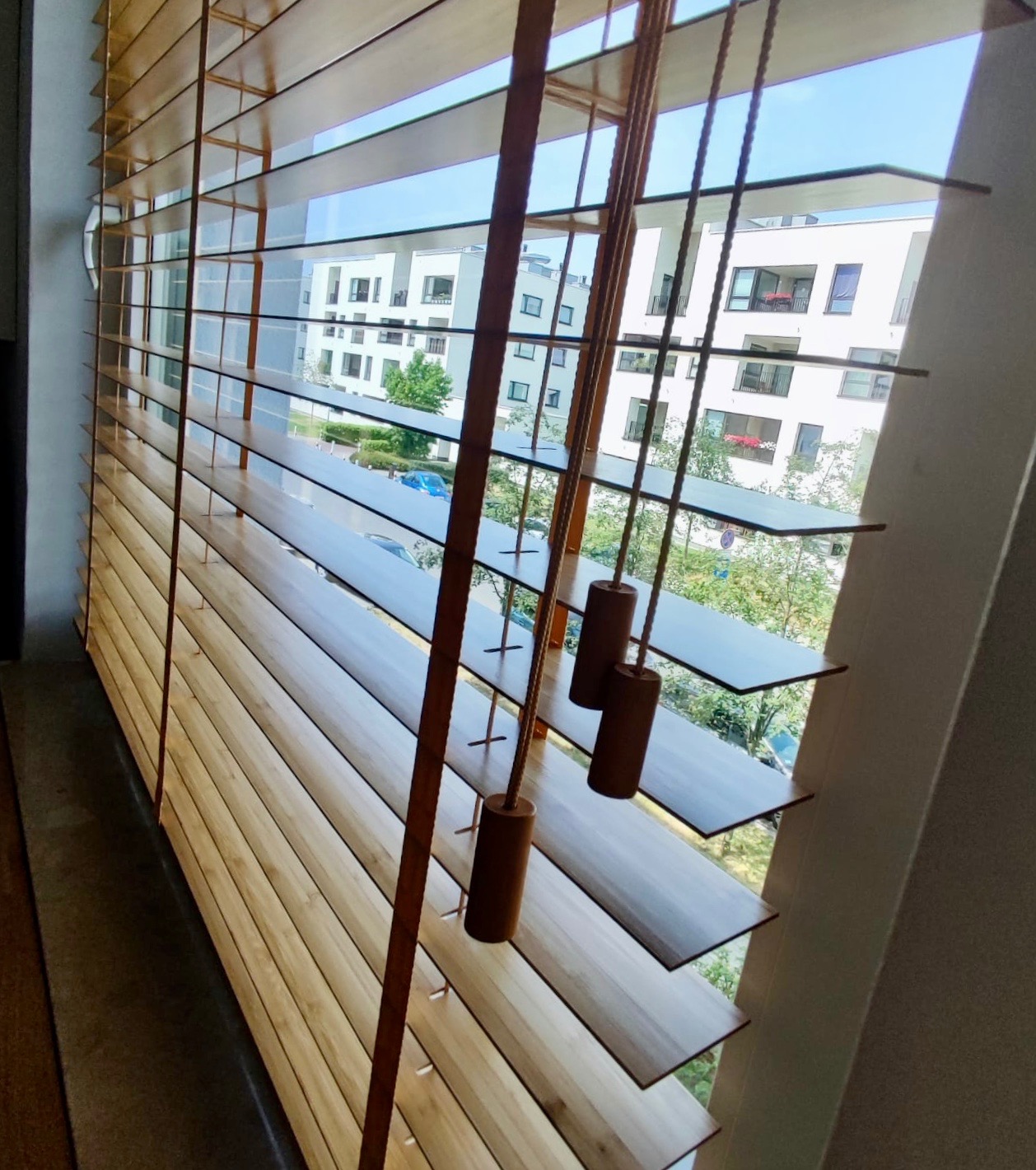 Drewniane listwy okienne w kolorze jasnego drewna, z widokiem na nowoczesne budynki mieszkalne w tle, sznurki do regulacji z drewnianymi obciążnikami.