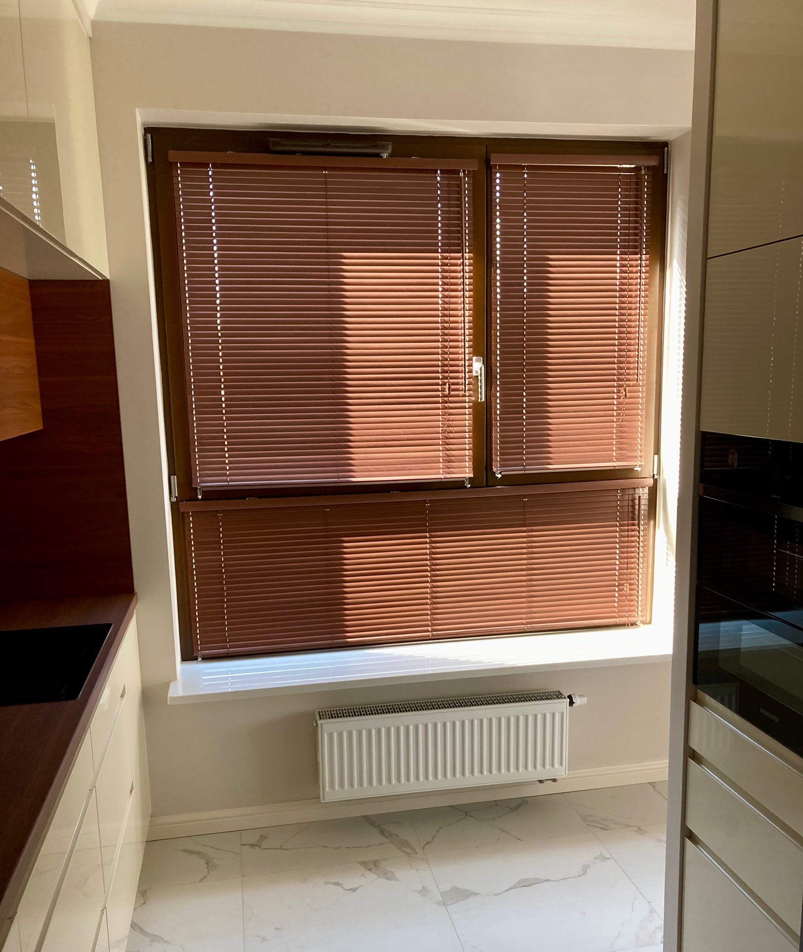 Brązowe poziome osłony okienne w nowoczesnej kuchni z marmurową podłogą, częściowo zasłonięte promieniami słonecznymi.