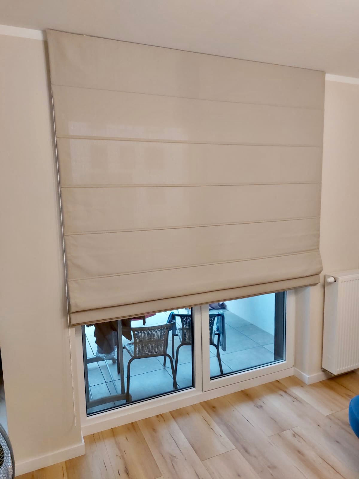 Beżowa, płaska osłona okienna w stylu rzymskim, częściowo opuszczona, widok na balkon z meblami rattanowymi, jasne wnętrze z drewnianą podłogą.
