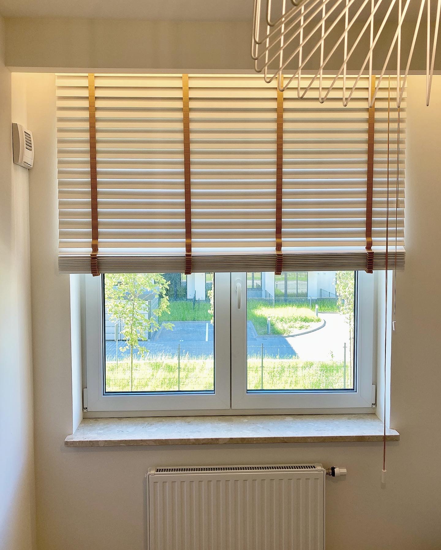 Jasne, poziome osłony okienne z brązowymi taśmami podtrzymującymi, widok na zieleń na zewnątrz, dekoracyjna lampa sufitowa w tle.