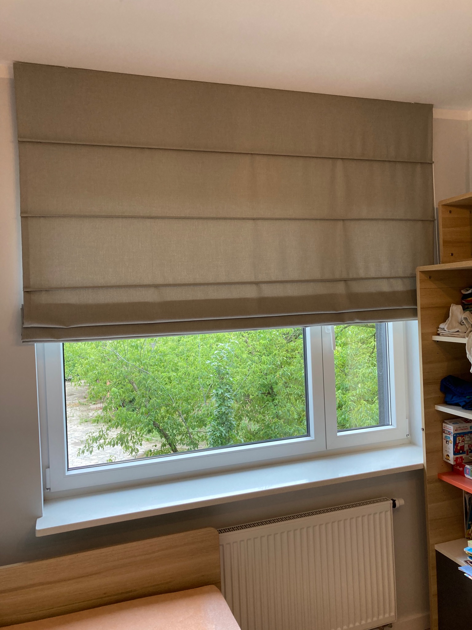 Beżowa tekstylna osłona okienna w stylu rzymskim, częściowo zasłonięta, widok na zieleń za oknem, grzejnik pod parapetem.