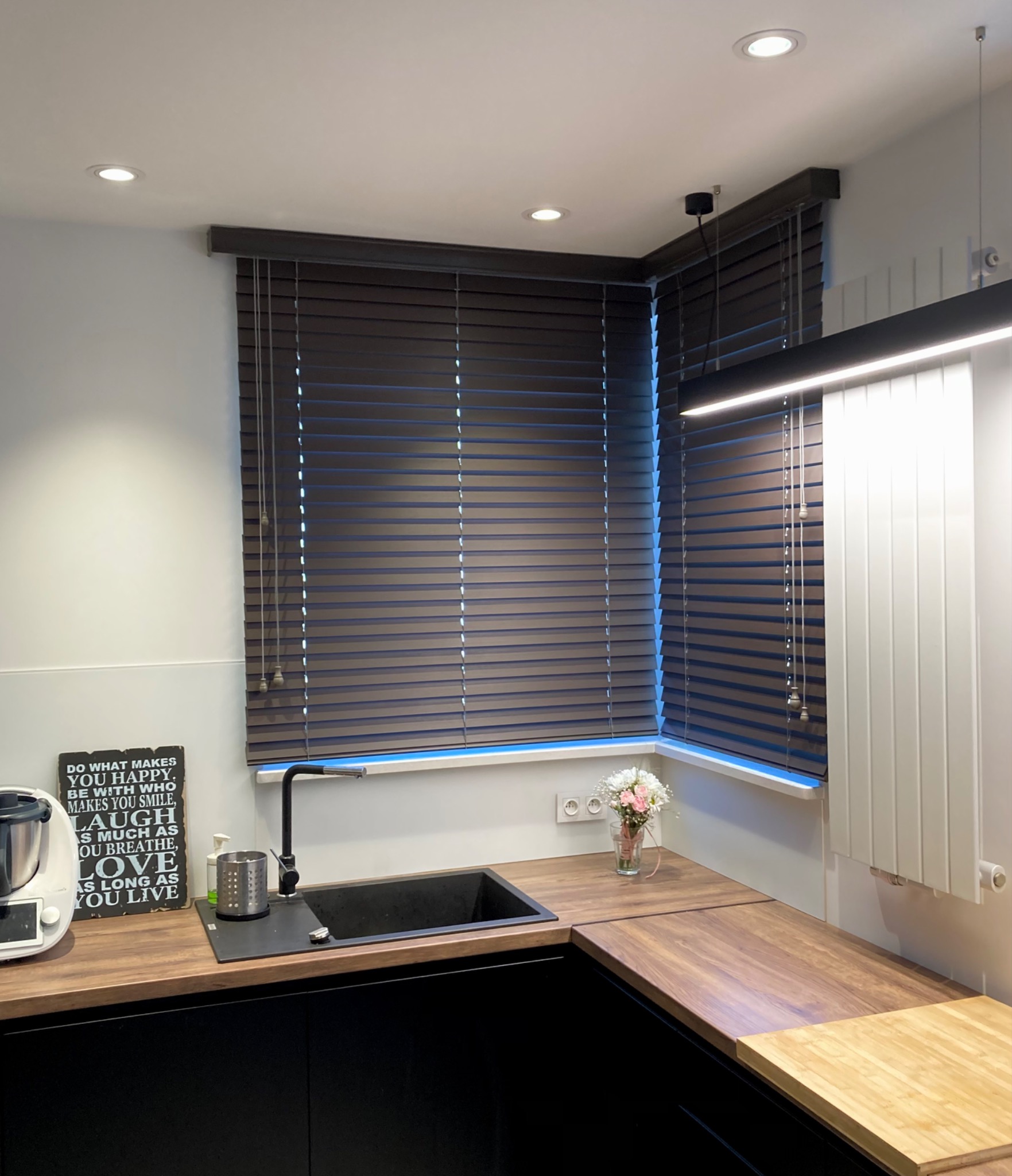 Ciemnobrązowe żaluzje drewniane w nowoczesnej kuchni z czarnymi szafkami i drewnianym blatem, oświetlone światłem punktowym i lampą wiszącą.
