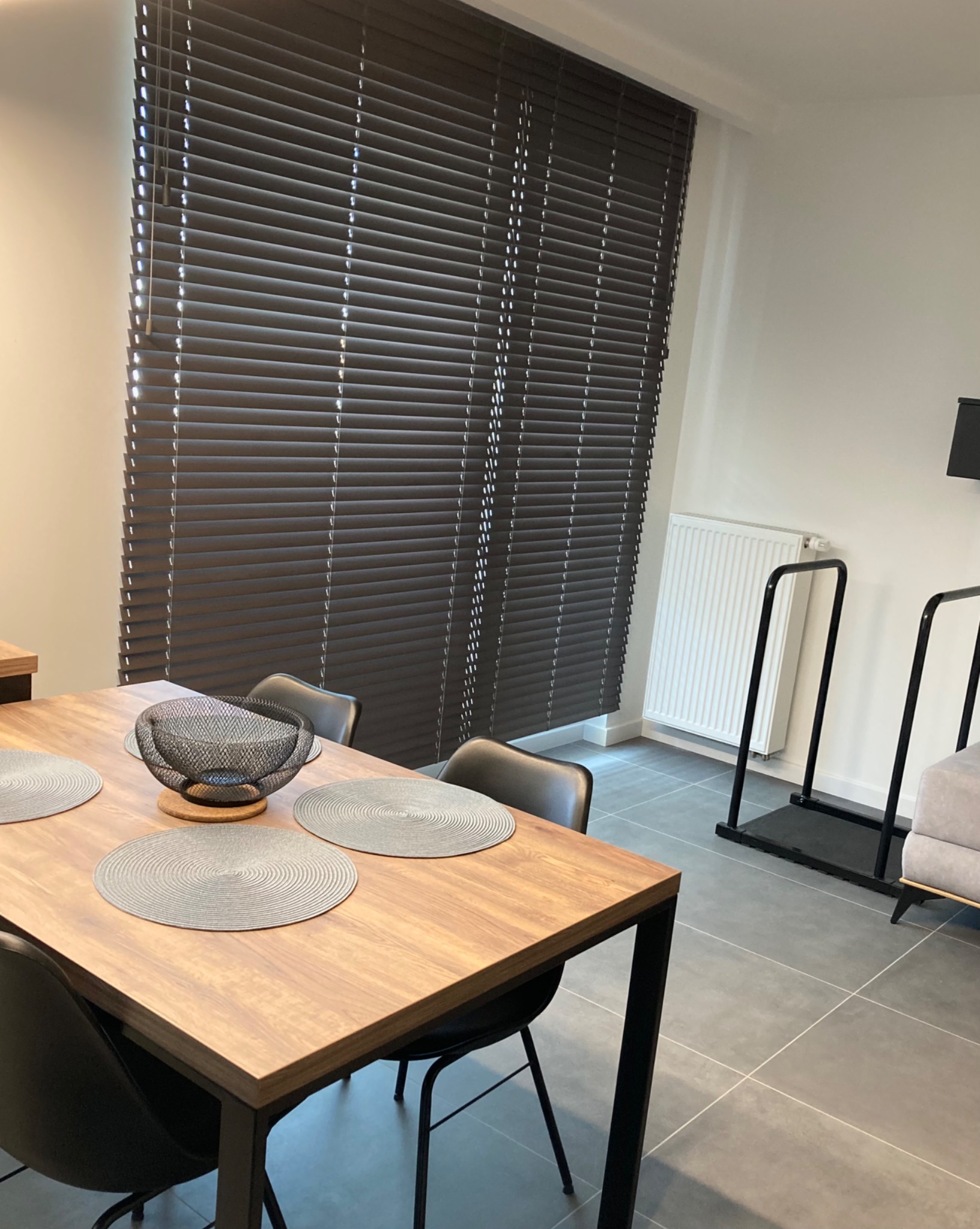 Brązowe poziome osłony okienne w nowoczesnym wnętrzu jadalni z drewnianym stołem i czarnymi krzesłami.