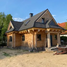 Nowo wybudowany, jasny, parterowy budynek z bali z ciemną dachówką, widoczne okna dachowe, drewniana drabina przy wejściu i przyczepka na podwórku.