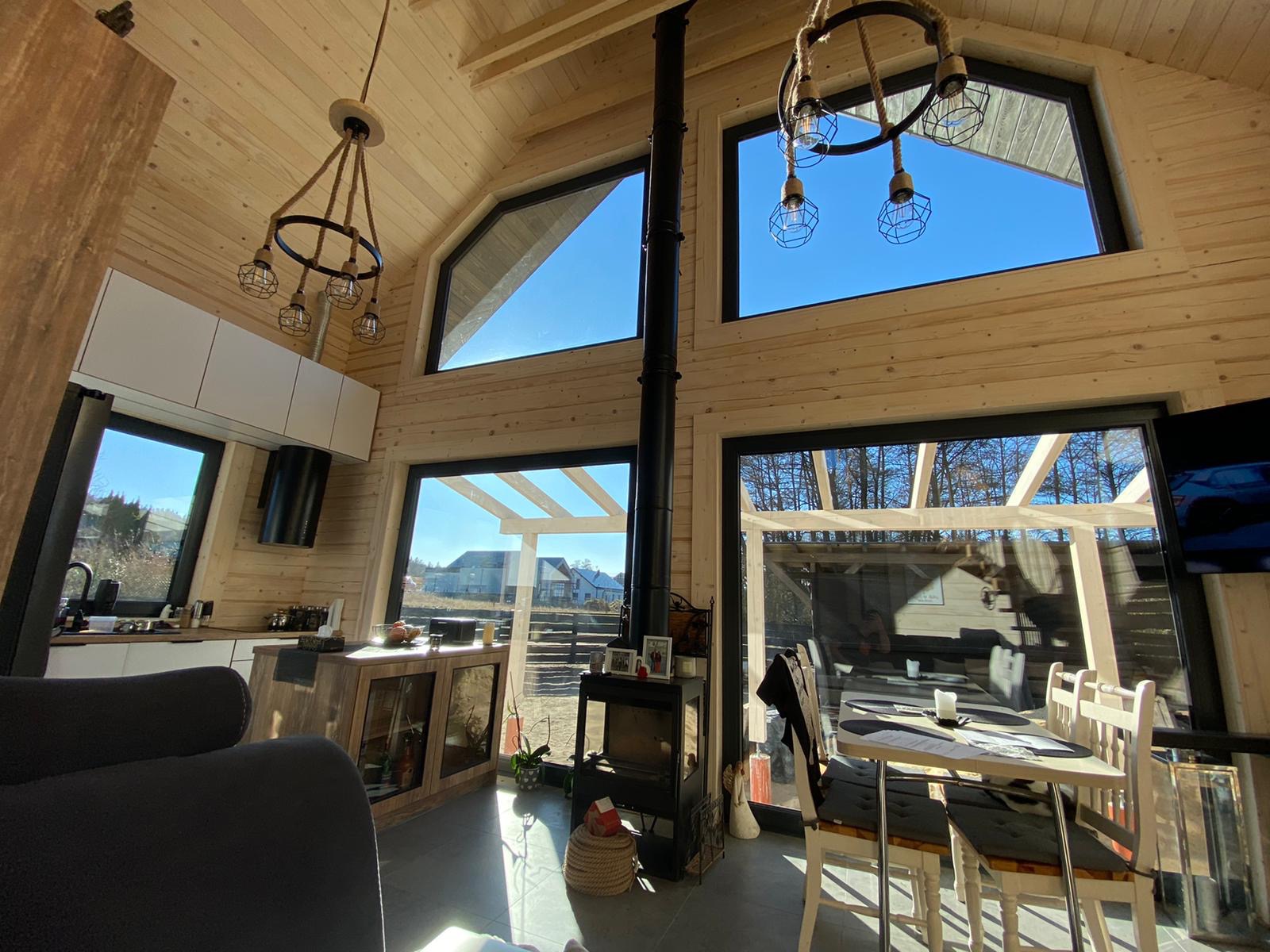 Jasne wnętrze domu z bali z wysokim sufitem, oknami o nietypowych kształtach i kominkiem, widok na zewnątrz na zabudowę jednorodzinną.