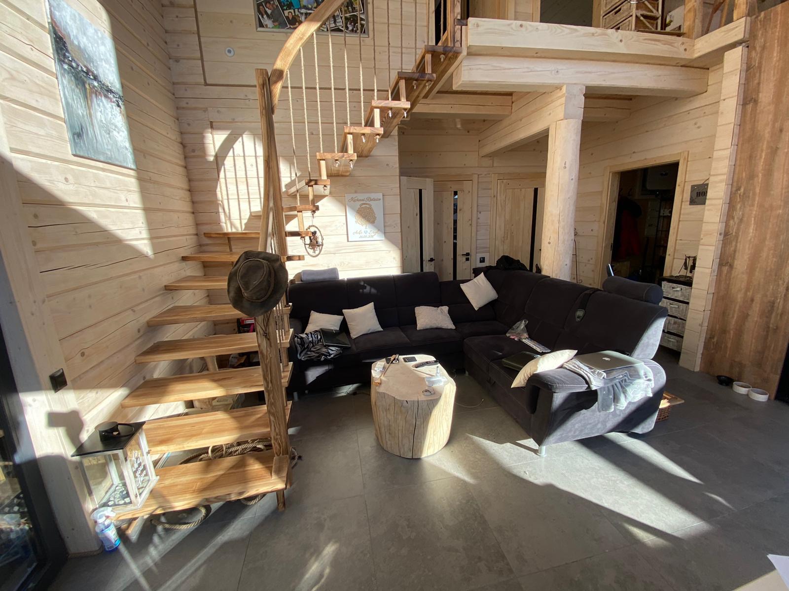 Wnętrze domu z drewnianych bali z drewnianymi schodami z linową barierką, antresolą i ciemną sofą narożną.