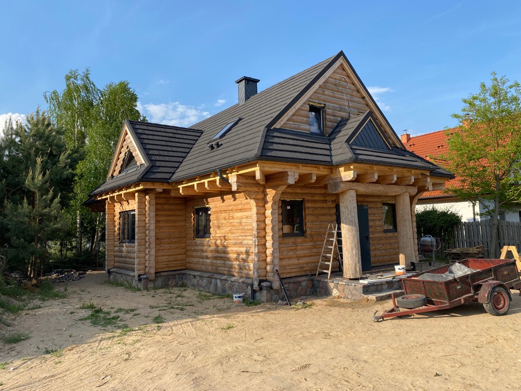 Nowo wybudowany, jasny, parterowy budynek z bali z ciemną dachówką, widoczne okna dachowe, drewniana drabina przy wejściu i przyczepka na podwórku.
