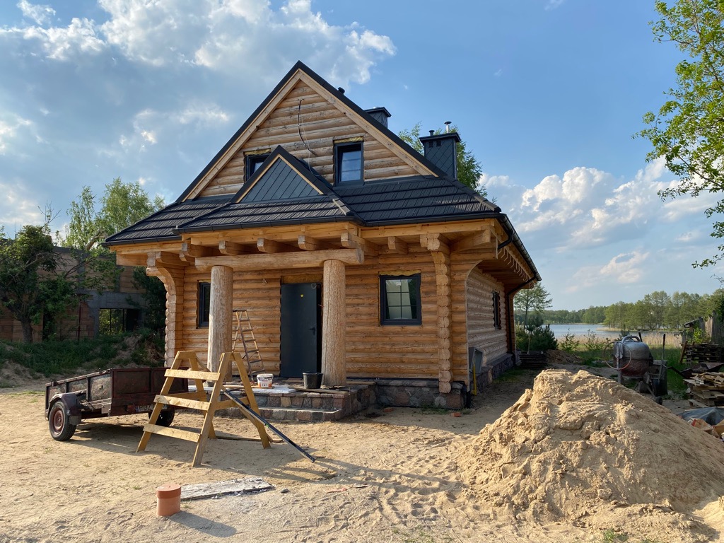 Surowy, parterowy budynek z bali z ciemną stolarką okienną i drzwiami, z widoczną przyczepką i stertą piasku na placu budowy w słoneczny dzień, z jeziorem w tle.