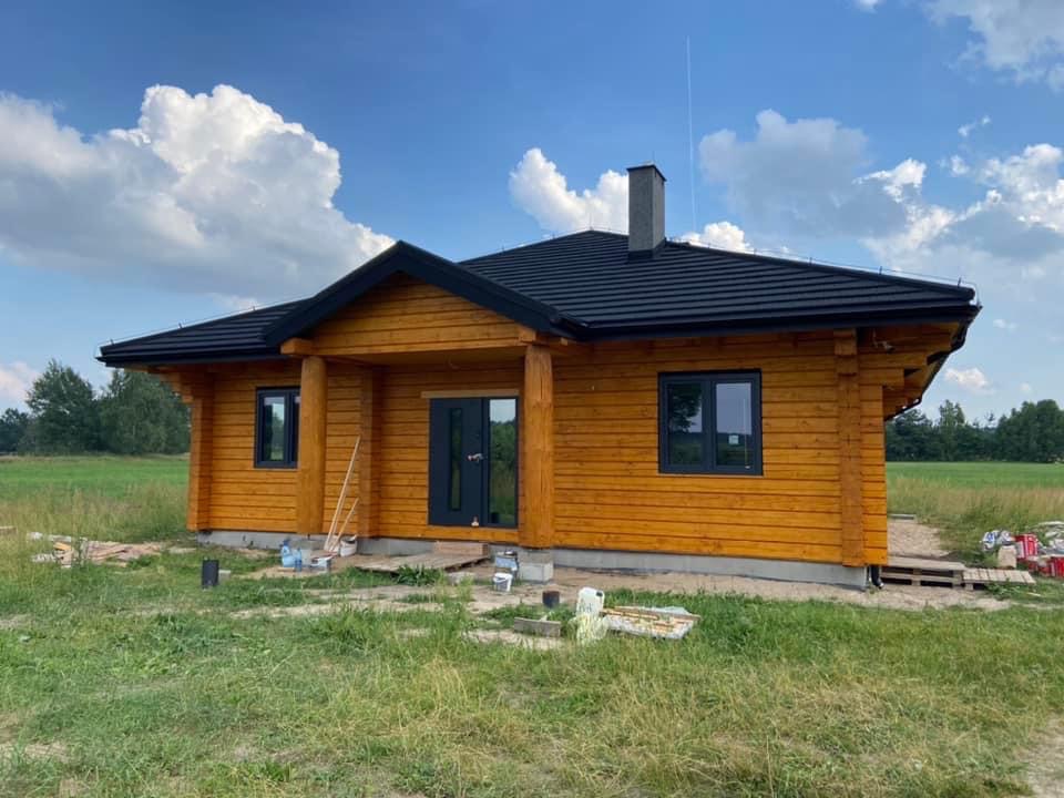 Nowoczesny, parterowy dom z drewnianych bali z czarnym dachem i stolarką okienną, usytuowany na zielonej działce pod błękitnym niebem z chmurami.