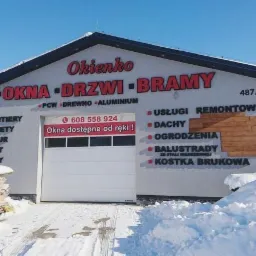 Budynek firmy Okienko z reklamą okien, drzwi i bram, z widoczną bramą garażową, ośnieżony teren przed budynkiem, okna w śniegu.