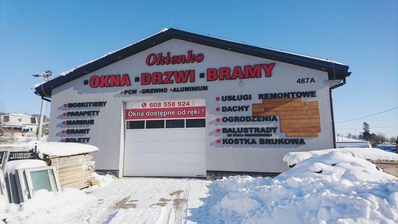 Budynek firmy Okienko z reklamą okien, drzwi i bram, z widoczną bramą garażową, ośnieżony teren przed budynkiem, okna w śniegu.