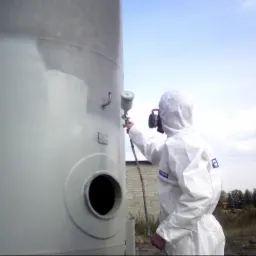 Pracownik w kombinezonie ochronnym natryskuje farbę na duży metalowy zbiornik, widoczny fragment pomalowanej powierzchni i pistolet lakierniczy.