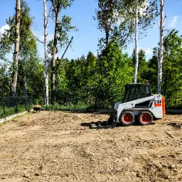 Kompaktowa ładowarka Bobcat S100 na wyrównanym, przygotowanym terenie z widocznymi śladami pracy, w tle drzewa liściaste i ogrodzenie siatkowe.