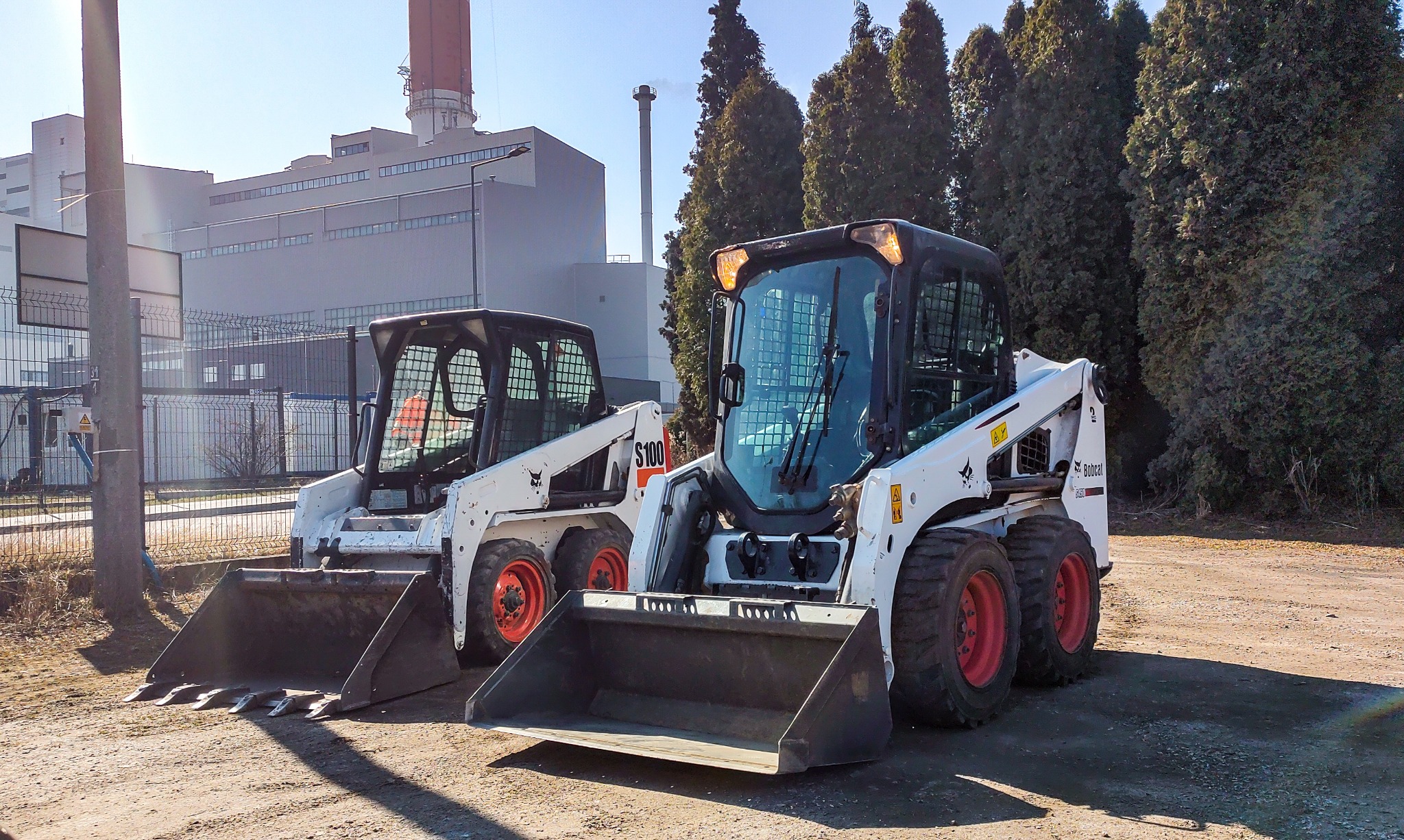 Dwa białe miniładowarki Bobcat zaparkowane na placu budowy, z fabryką w tle i wysokim kominem.