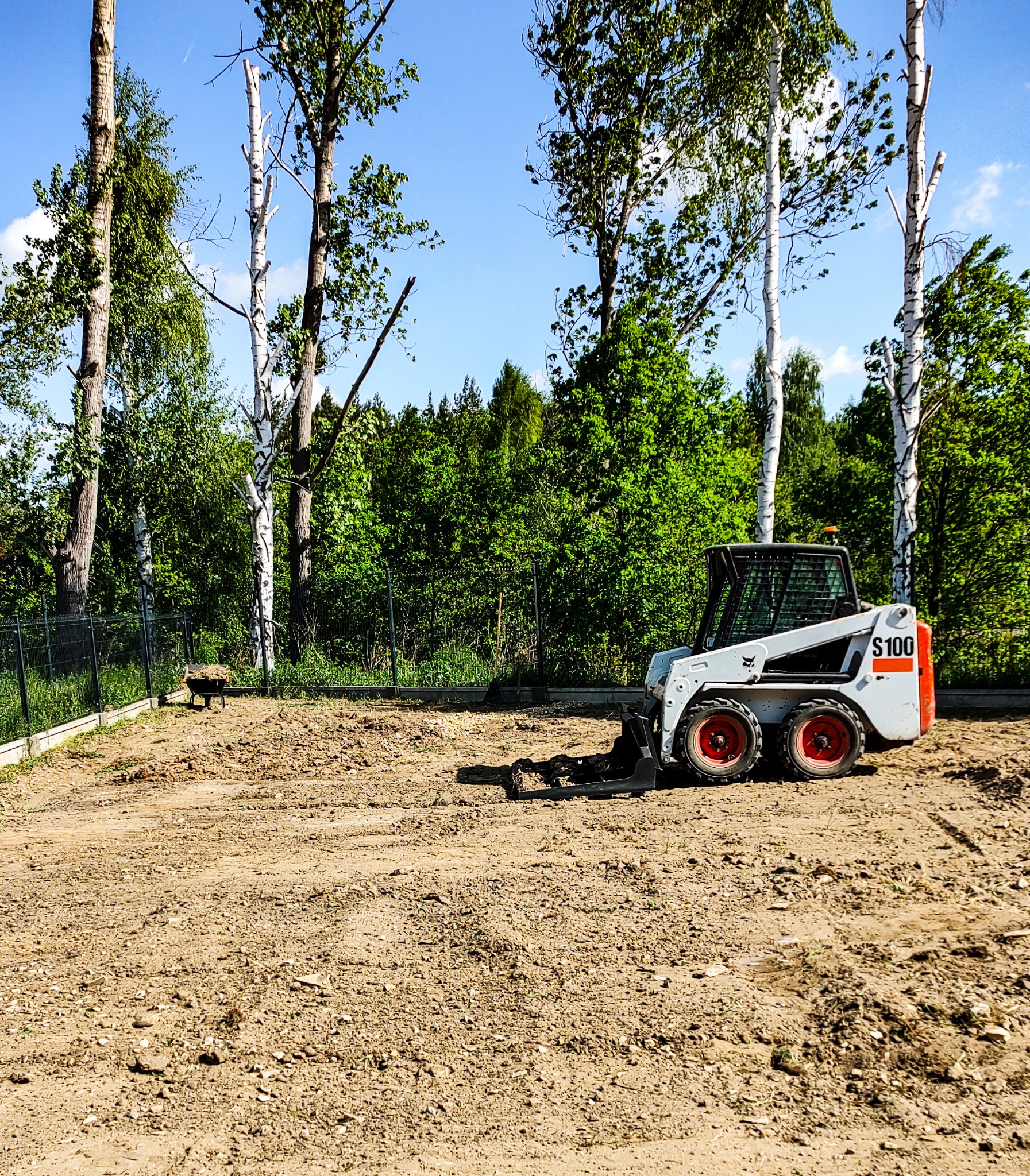 Kompaktowa ładowarka Bobcat S100 na wyrównanym, przygotowanym terenie z widocznymi śladami pracy, w tle drzewa liściaste i ogrodzenie siatkowe.