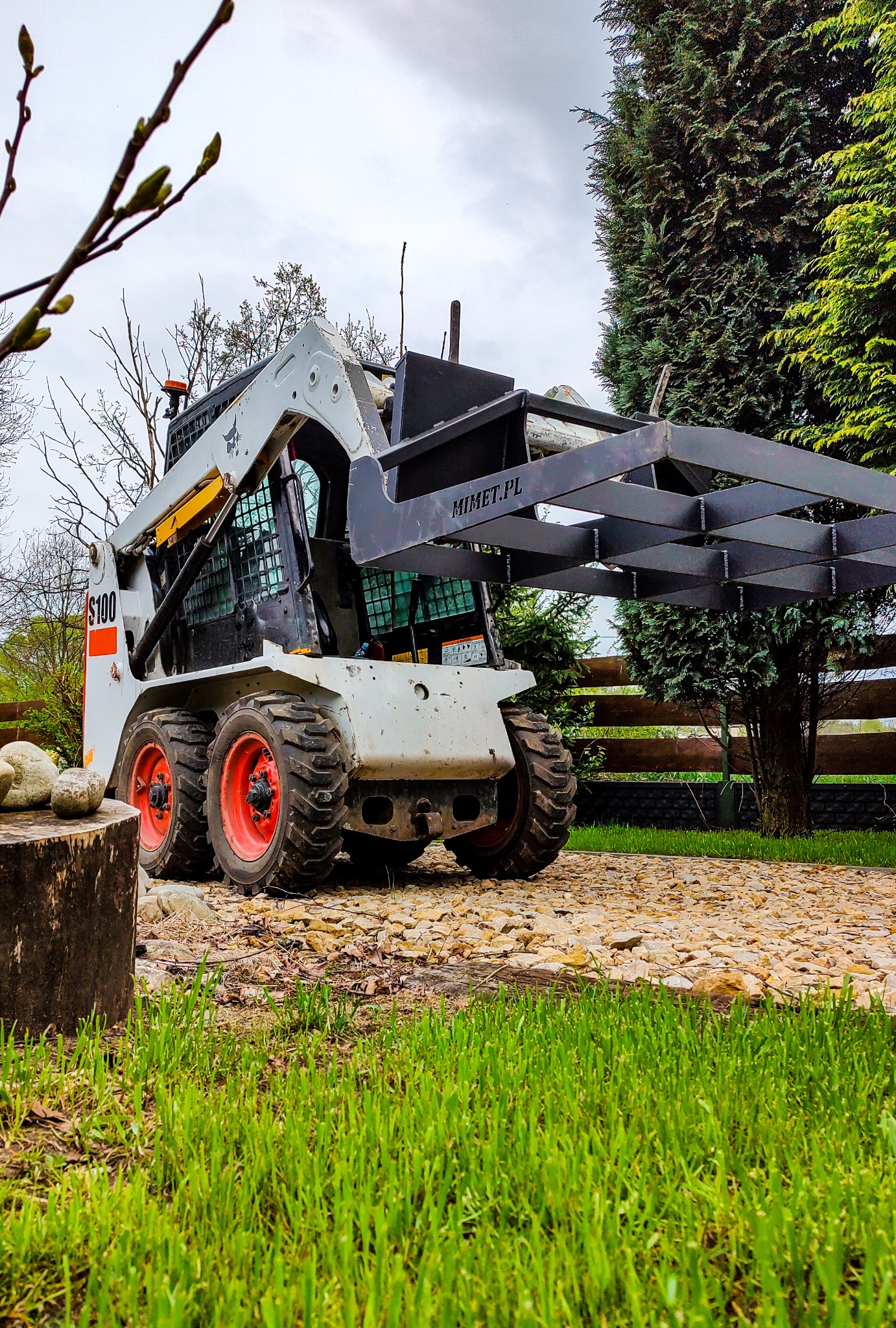Bobcat S100 z zamontowanym chwytakiem do drzewa na nierównym terenie z kamieniami i trawą, widoczne drzewa w tle.