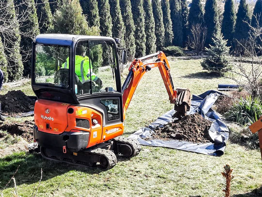 Pomarańczowa mini koparka Kubota z operatorem w żółtej kamizelce nabiera ziemię na czarną folię rozłożoną na trawniku, w tle rząd tuj i mała choinka.