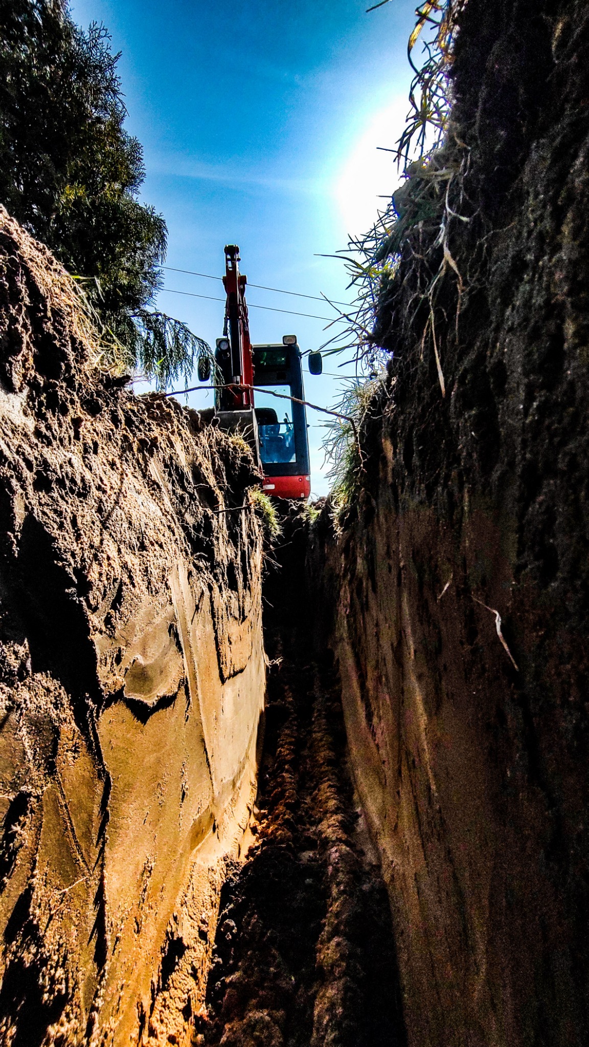 Wąski wykop w ziemi widziany z dołu, z małą koparką na końcu wykopu, pod błękitnym niebem.