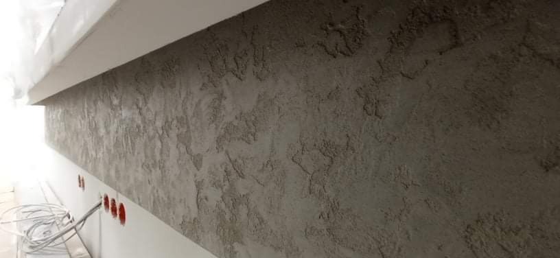 Ściana pokryta szarą, dekoracyjną strukturą z widocznymi nierównościami i wzorem, z przygotowanymi otworami na gniazdka elektryczne i wystającymi kablami.