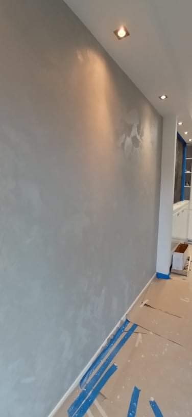 Ściana z efektem dekoracyjnym w odcieniach szarości, zabezpieczona niebieską taśmą malarską przy podłodze, oświetlona punktowymi lampami sufitowymi.