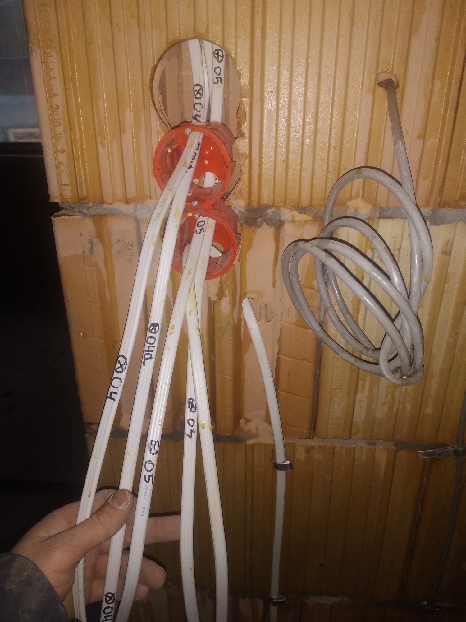 Instalacja elektryczna w trakcie montażu: białe przewody z oznaczeniami wchodzą do pomarańczowych puszek instalacyjnych osadzonych w ceglanej ścianie. Widoczne również zwinięte szare kable i ręka...