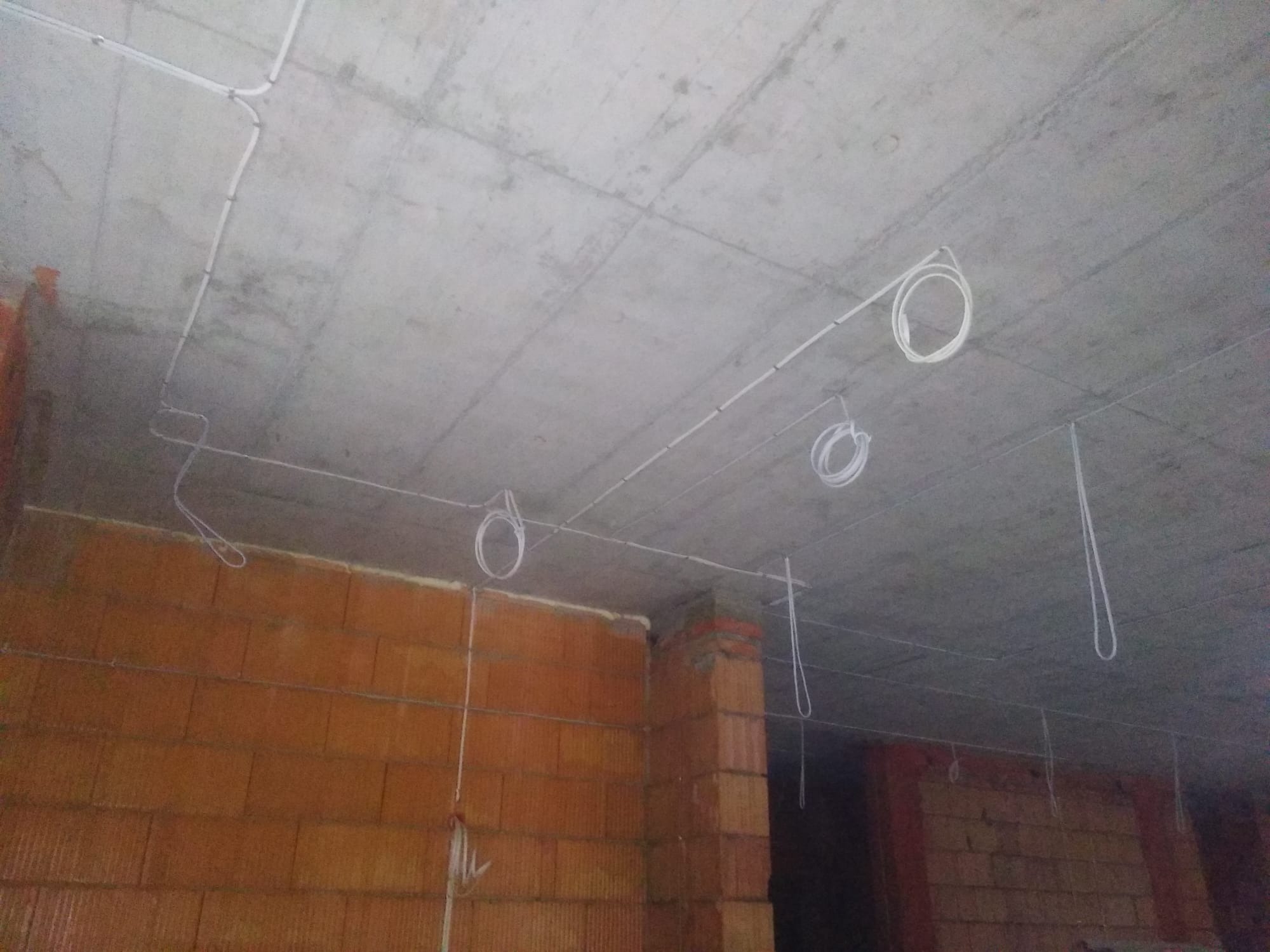 Instalacja elektryczna natynkowa na surowym, betonowym suficie w trakcie budowy, z widocznymi ceglanymi ścianami i zwisającymi przewodami.
