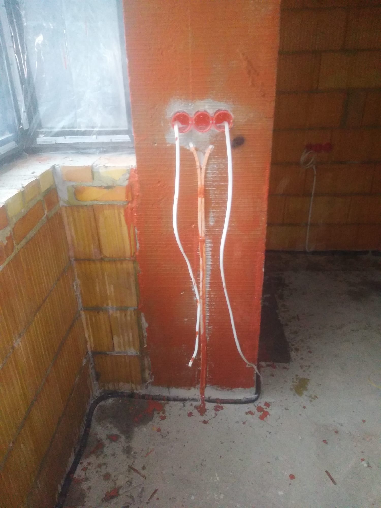 Instalacja elektryczna podtynkowa w trakcie montażu, widoczne kable w peszlach wychodzące z puszki w ścianie z cegły, obok okno z folią zabezpieczającą.