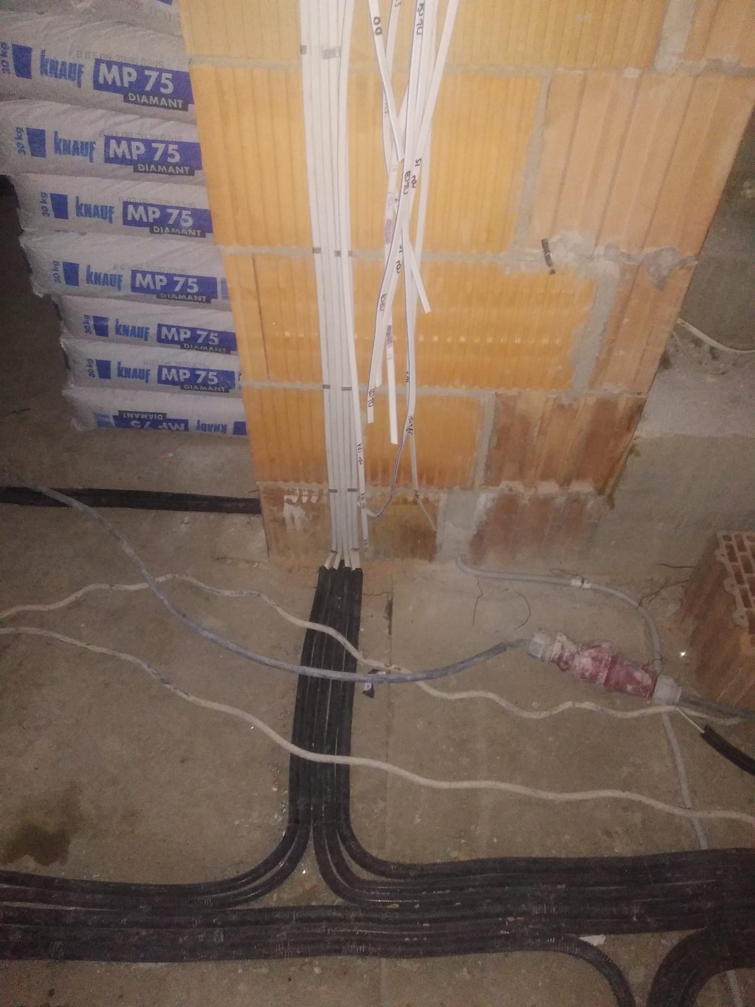 Instalacja elektryczna w trakcie montażu: wiązki kabli prowadzone w ścianie z cegły, widoczne worki z zaprawą murarską w tle, kable ułożone na podłodze.