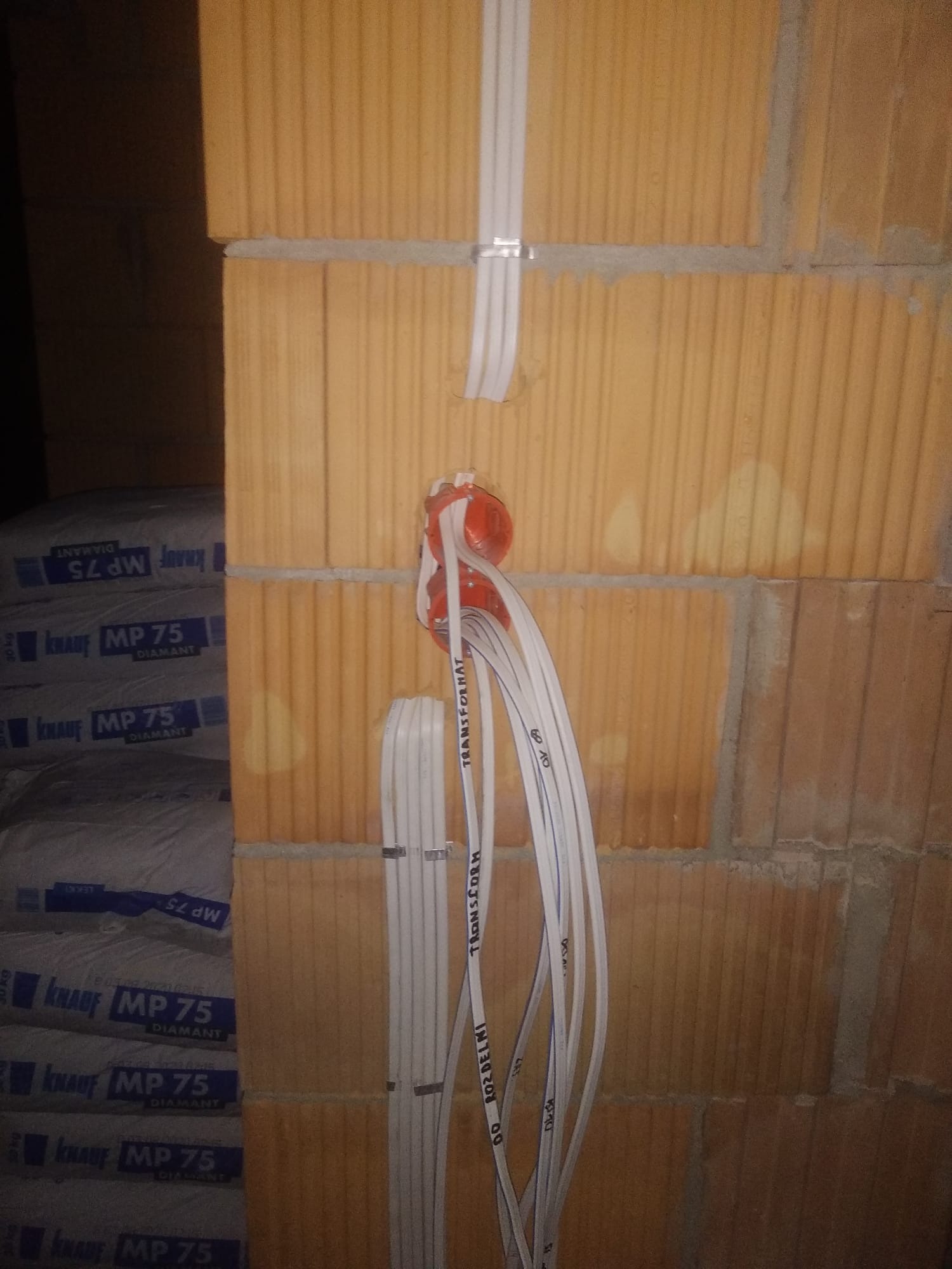 Instalacja elektryczna w trakcie montażu w ścianie z cegły dziurawki. Widoczne przewody w peszlach wychodzące z puszki instalacyjnej, przymocowane klamrami do ściany. W tle worki z zaprawą murarską...