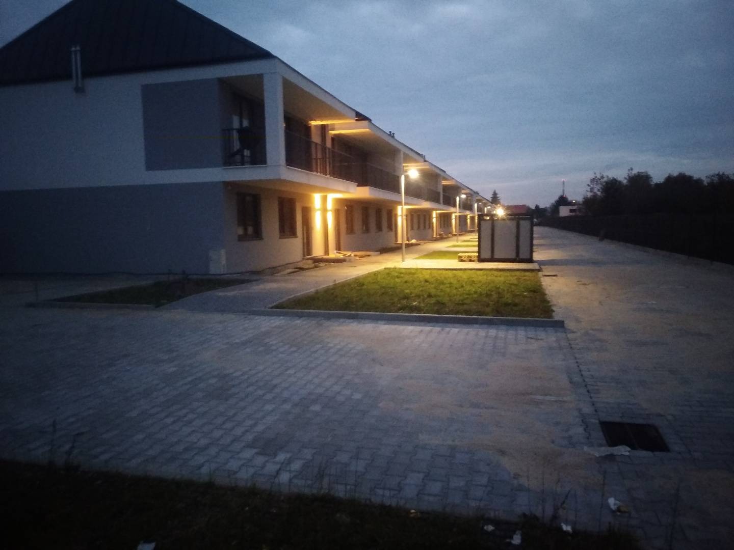 Nowe budynki mieszkalne w trakcie lub tuż po zakończeniu prac instalacyjnych oświetlenia zewnętrznego, widoczne lampy uliczne wzdłuż chodnika i oświetlone balkony.