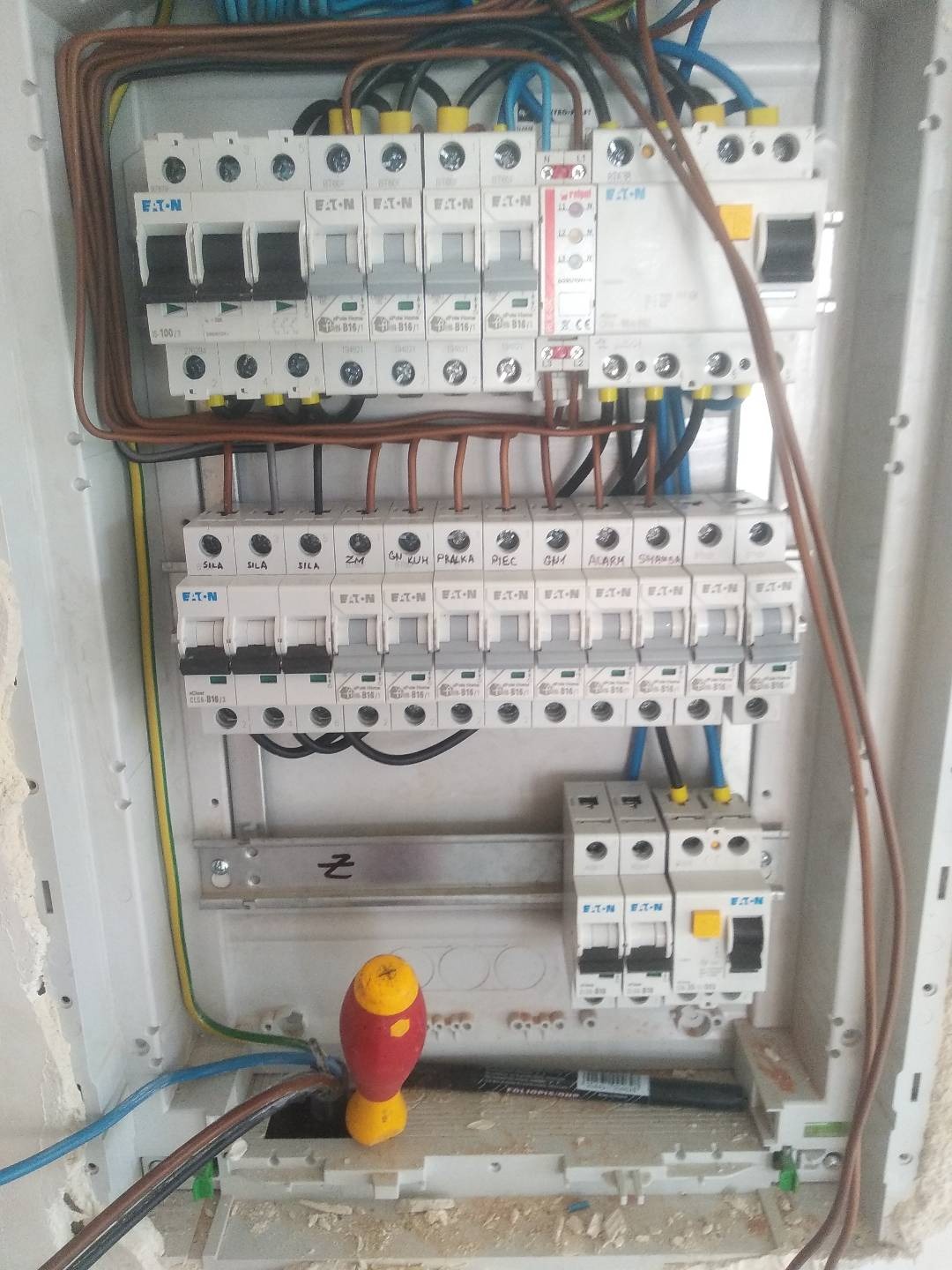 Rozdzielnica elektryczna marki Eaton z widocznymi bezpiecznikami i przewodami, obok leży czerwono-żółty śrubokręt i czarny marker.