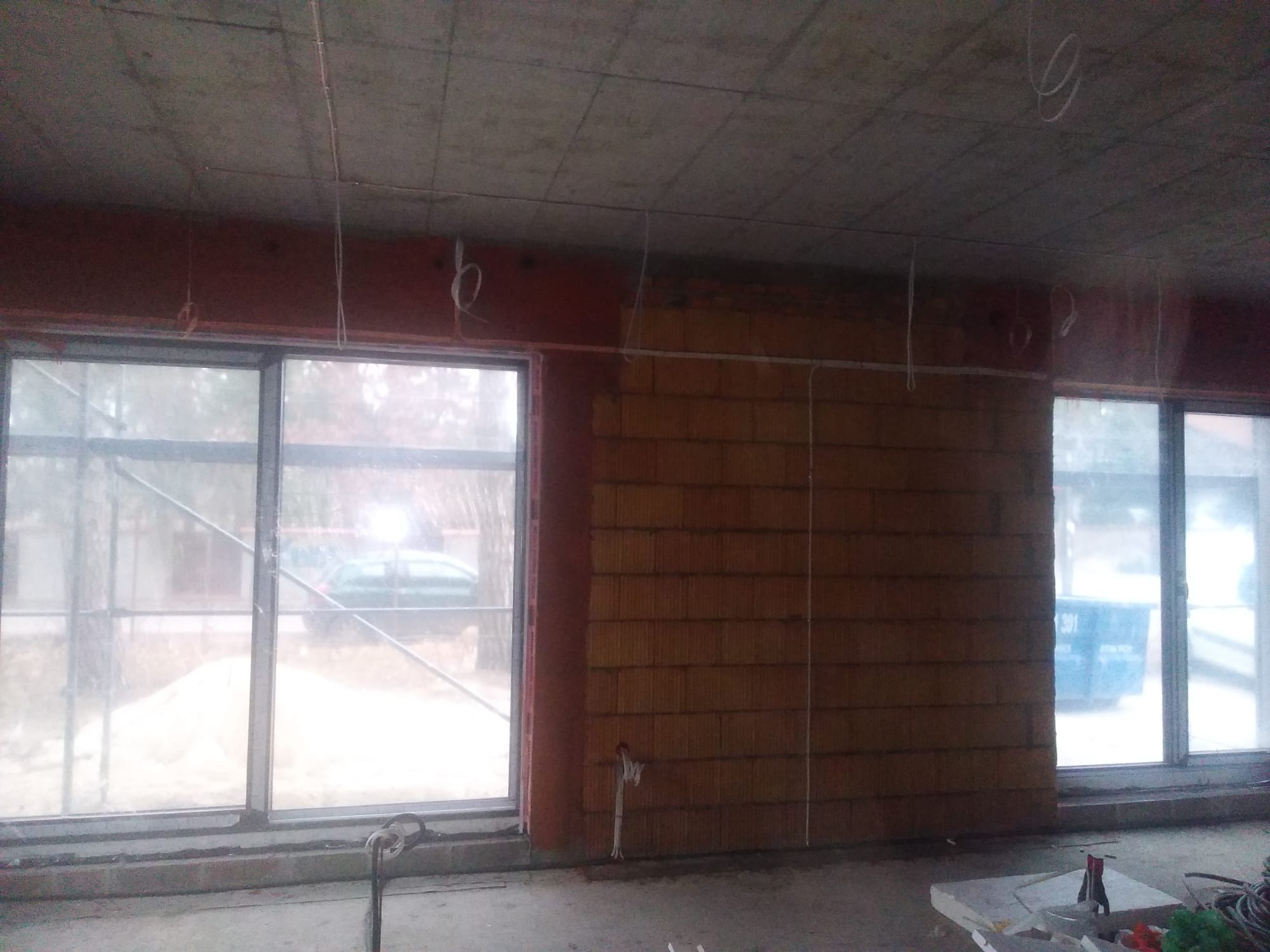 Wnętrze w stanie surowym z widocznymi przewodami elektrycznymi zwisającymi z sufitu, ściany z cegły dziurawki, okna z widokiem na zewnątrz, oraz narzędzia na podłodze.