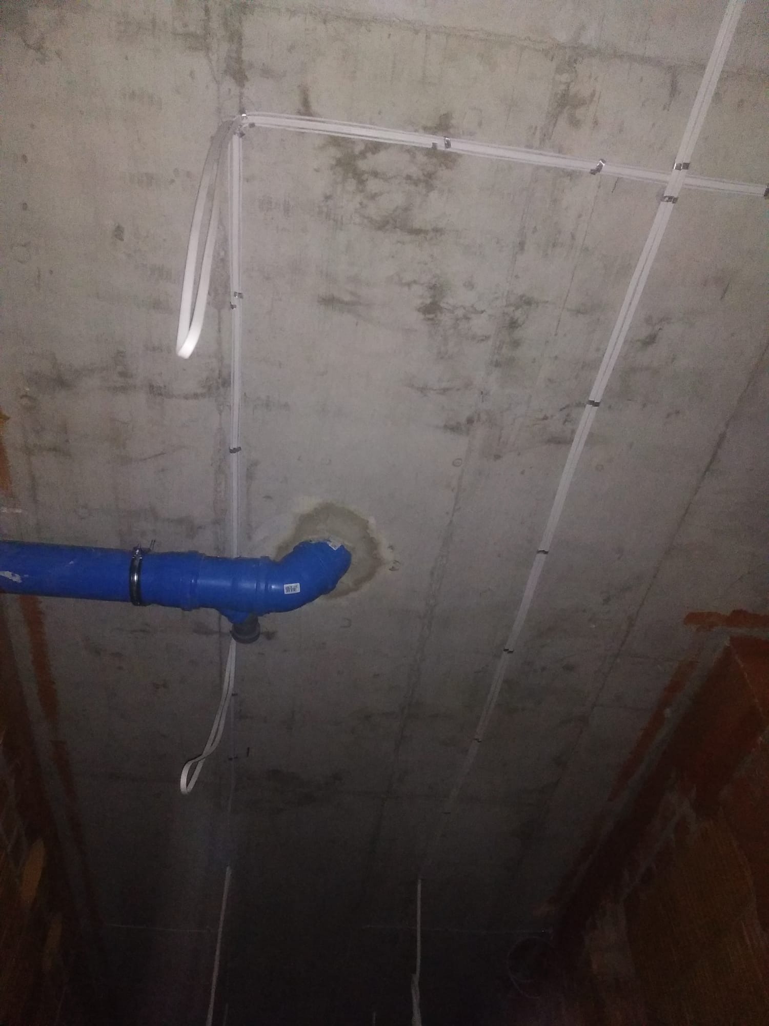 Surowy betonowy sufit z zamontowanymi białymi listwami instalacyjnymi i niebieską rurą kanalizacyjną z obejmą.