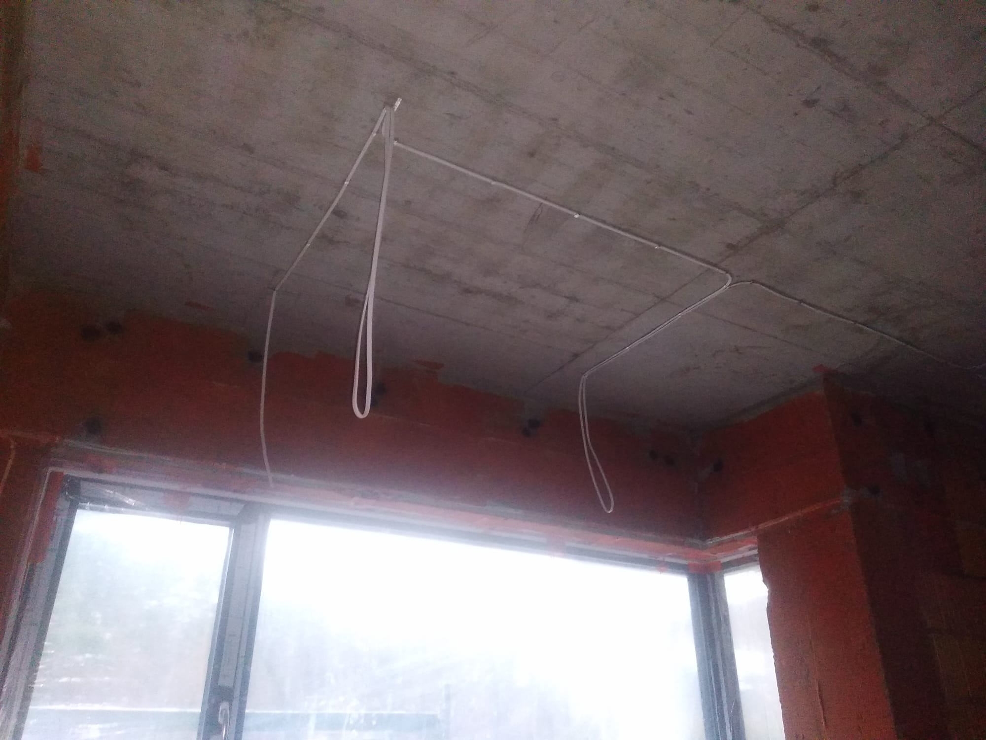 Surowy betonowy sufit z prowizorycznie zamontowanymi białymi kablami elektrycznymi, zwisającymi w pętlach, widok z wnętrza niedokończonego pomieszczenia z oknem.