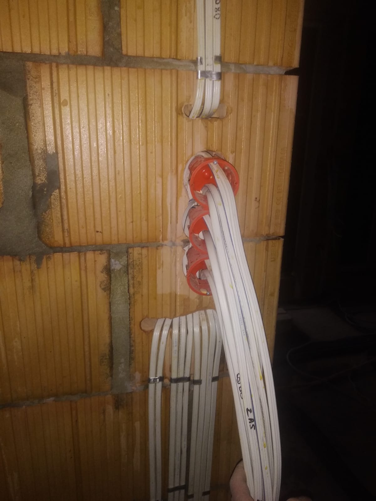 Kable elektryczne w białej osłonie, prowadzone w ścianie z pustaków ceramicznych, zabezpieczone metalowymi klamrami i wchodzące do pomarańczowych puszek instalacyjnych.