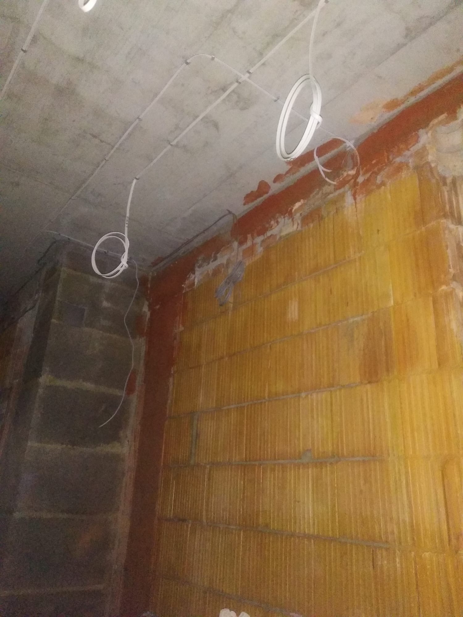 Surowe ściany z pustaków i betonowy sufit z widocznymi, częściowo zamontowanymi przewodami elektrycznymi, zwisającymi w pętlach.