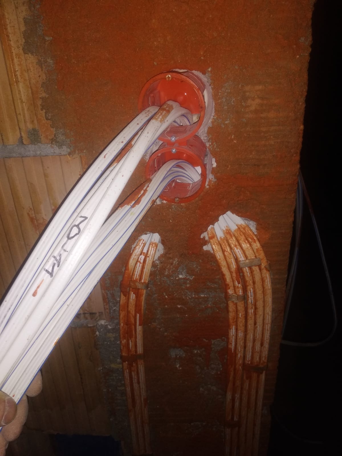 Instalacja elektryczna w trakcie montażu: białe kable z niebieskimi paskami wchodzą do pomarańczowych puszek osadzonych w czerwonej, porowatej ścianie. Na kablach widoczne napisy markerem.