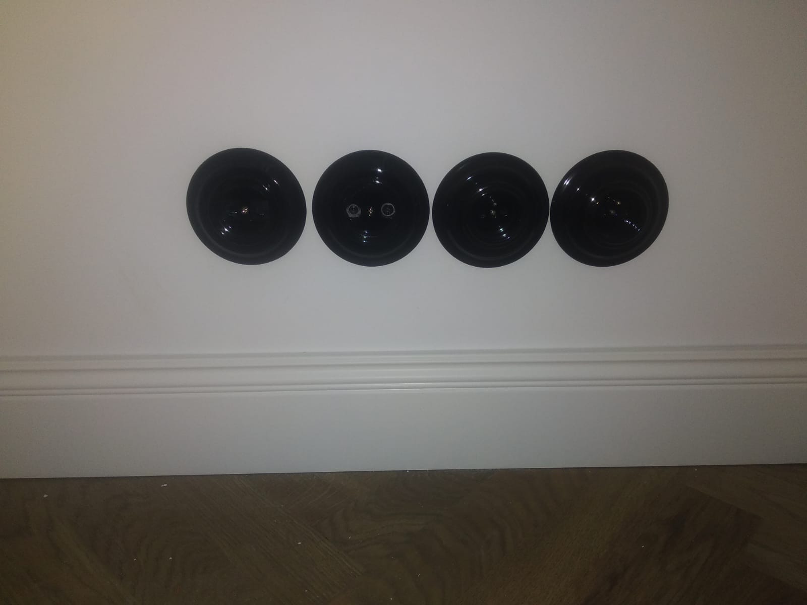 Cztery czarne, okrągłe gniazdka elektryczne zamontowane poziomo na białej ścianie nad białą listwą przypodłogową i drewnianą podłogą.
