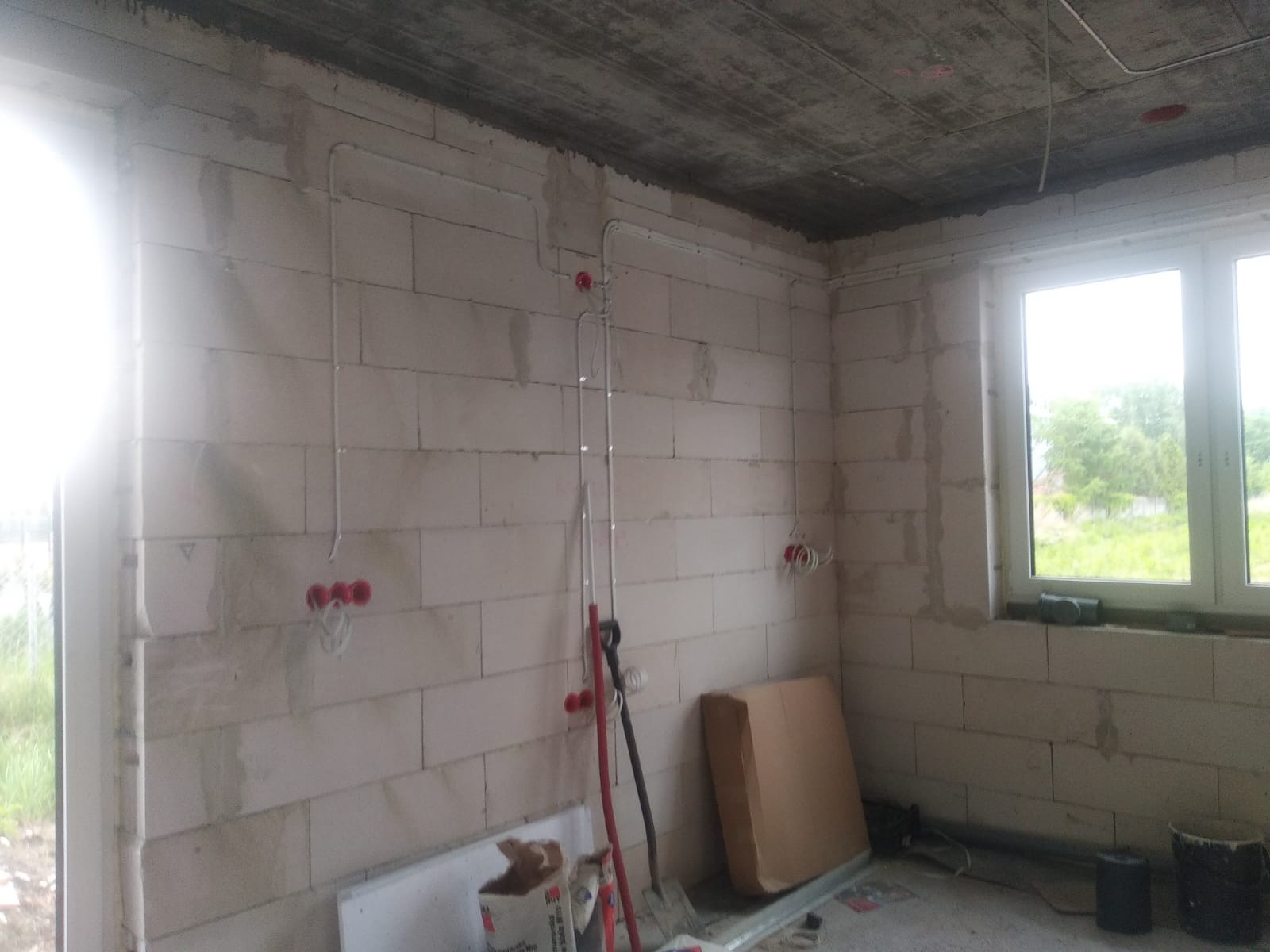 Surowy stan: Instalacja elektryczna w budynku z białych bloczków, widoczne przewody w peszlach, puszki podtynkowe, betonowy sufit, okno z widokiem na zieleń.
