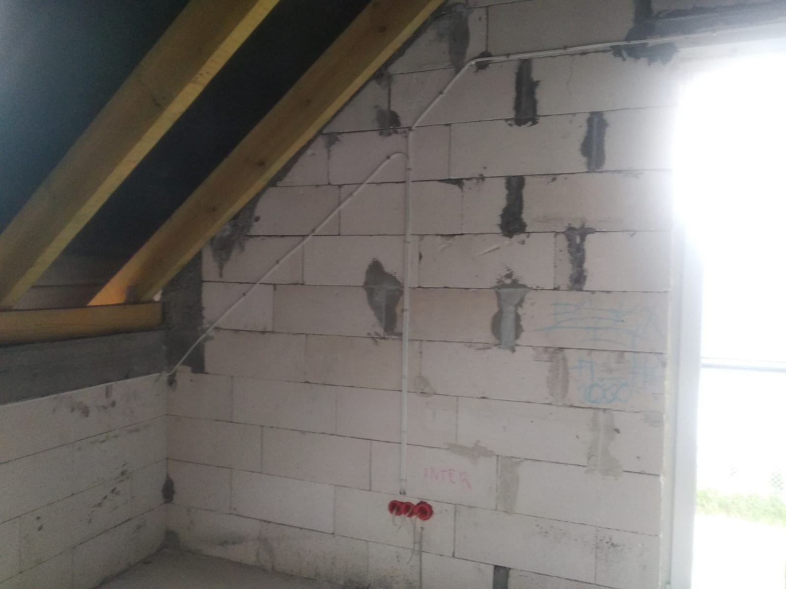 Instalacja elektryczna w surowym stanie poddasza: białe kable prowadzone po ścianie z bloczków, trzy czerwone gniazdka podtynkowego montażu, widoczne drewniane elementy konstrukcji dachu.