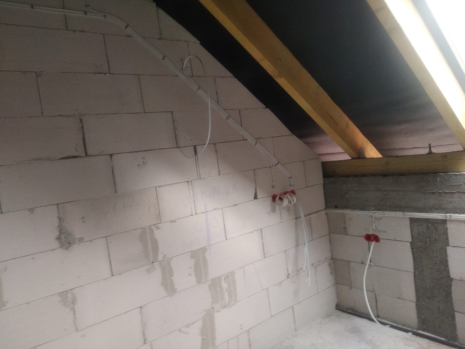 Instalacja elektryczna w surowym wnętrzu poddasza: przewody poprowadzone po ścianie z białych bloczków, widoczne puszki instalacyjne z wyprowadzonymi przewodami, drewniana konstrukcja dachu.